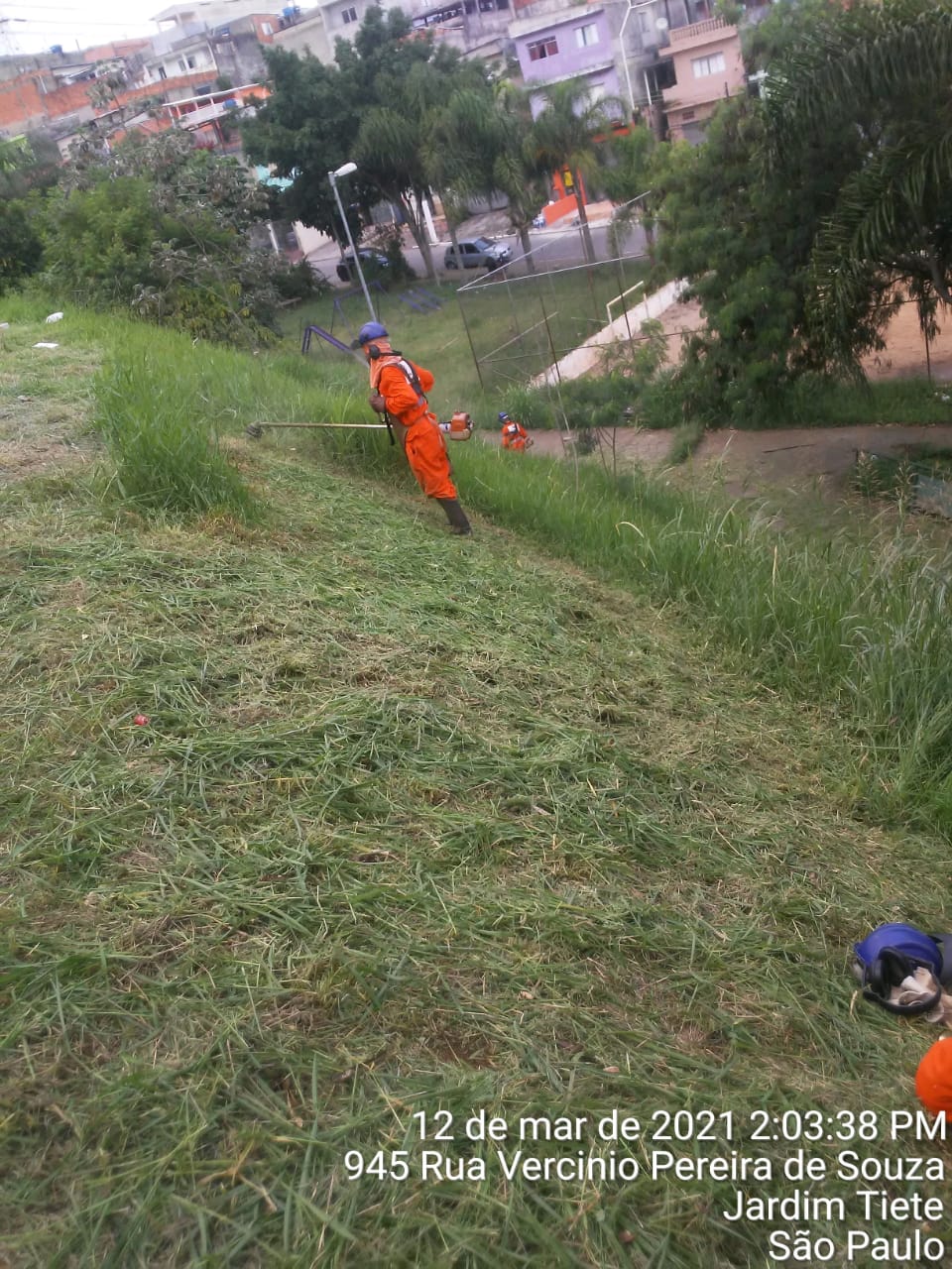 Dois servidores cortam grama em um terreno com desnível. O corpo está inclinado à esquerda. 